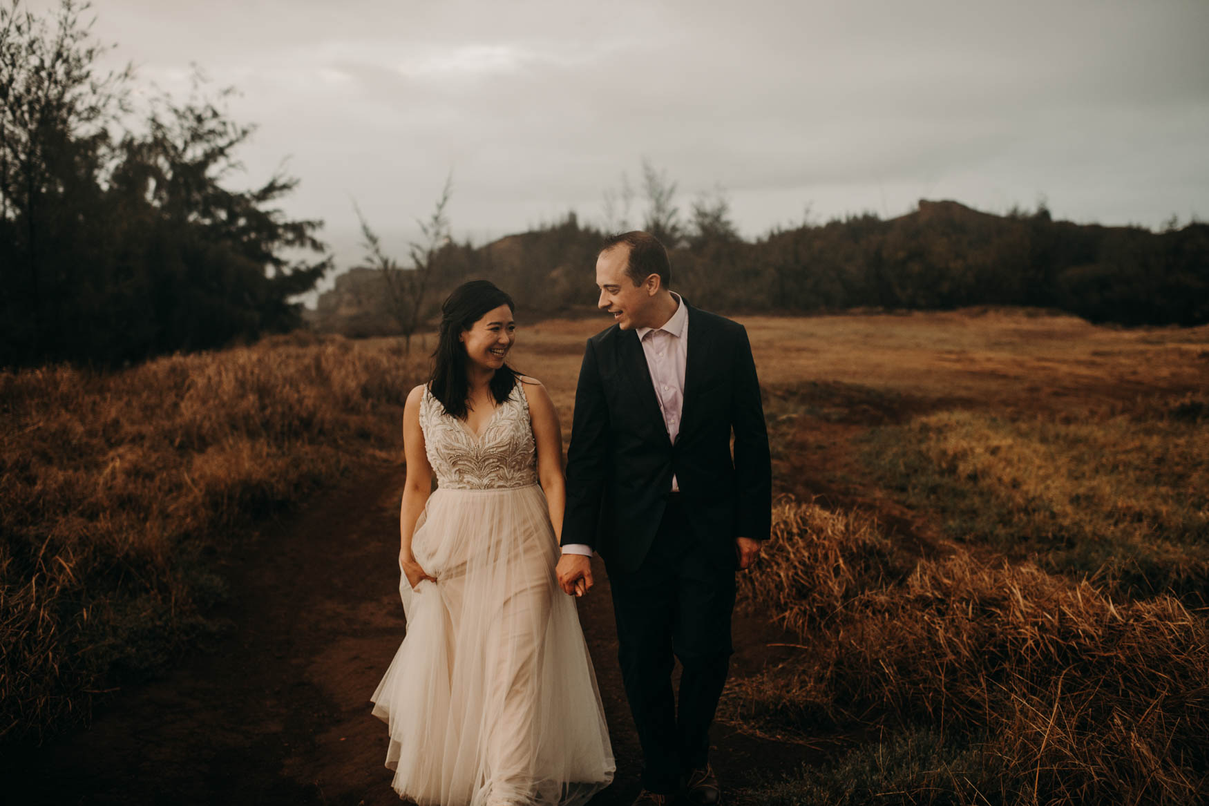 Andrew and Alison hold hands, looking at one another, smiling, as they walk toward the camera on a red clay path through tall yellow grass.