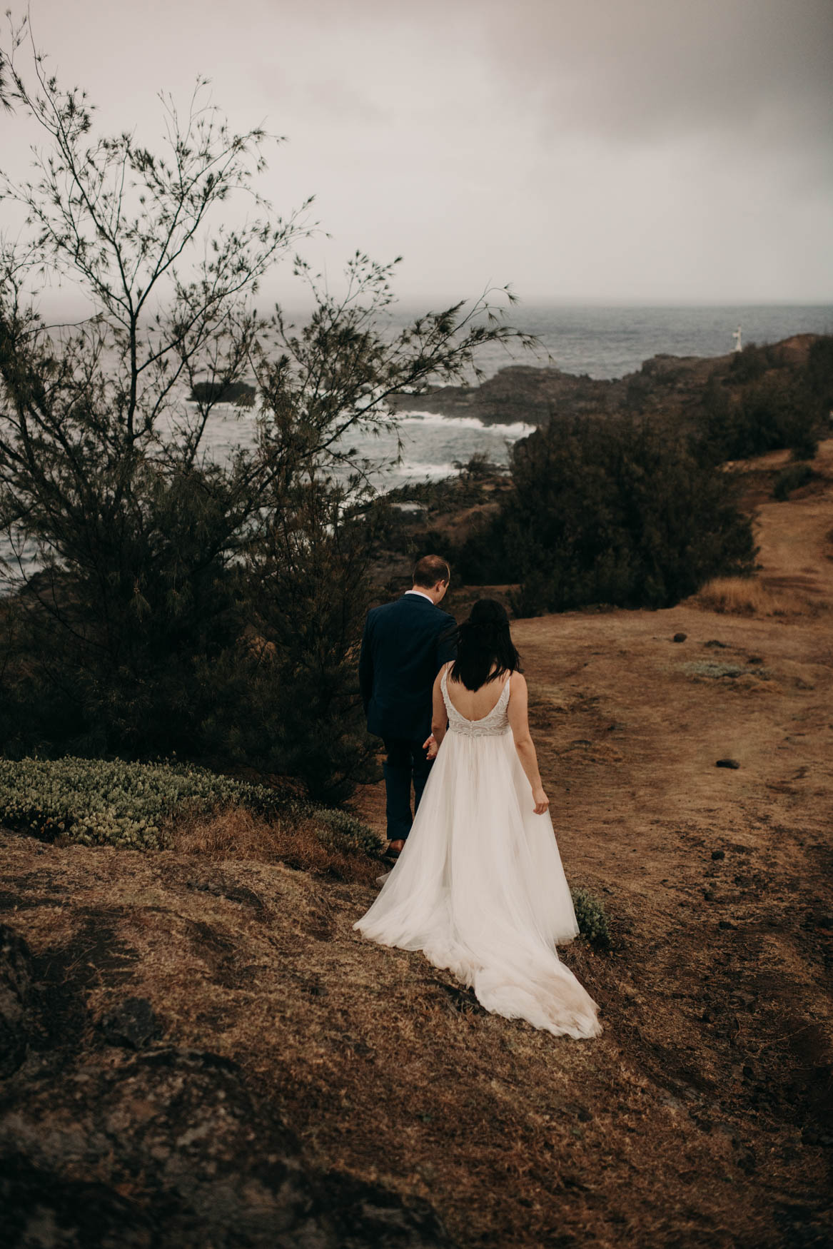 Andrew and Alison walk away from the camera, downhill, on a path with short yellow grass. Andrew is slightly ahead of Alison, holding her hand.