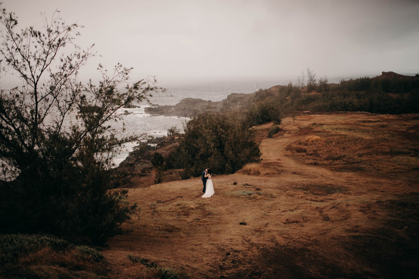 A distant shot with Andrew and Alison in the midframe. They stand in the mowed area of the field, cliffs and the ocean to their left, red clay pathways to their right. The hold eachother close and kiss.