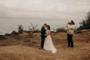 Andrew and Alison lean on one another, their forheads touching, while Euta blows the conch. The ocean and clouds behind them.