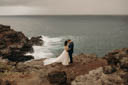 Andrew and Alison stand together on a cliff, the grey-blue ocean crashing on rocks behind and below them.