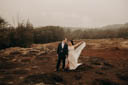 Andrew and Alison stand together in a red clay field with patches of yellow dry grass, mist rolling in, and green shrubs in the further background. They hold hands. Andrew looks at Alison, smiling. Alison looks at Andrew, smiling, while holding the train of her dress up with her left hand.