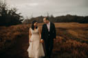 Andrew and Alison hold hands, looking at one another, smiling, as they walk toward the camera on a red clay path through tall yellow grass.
